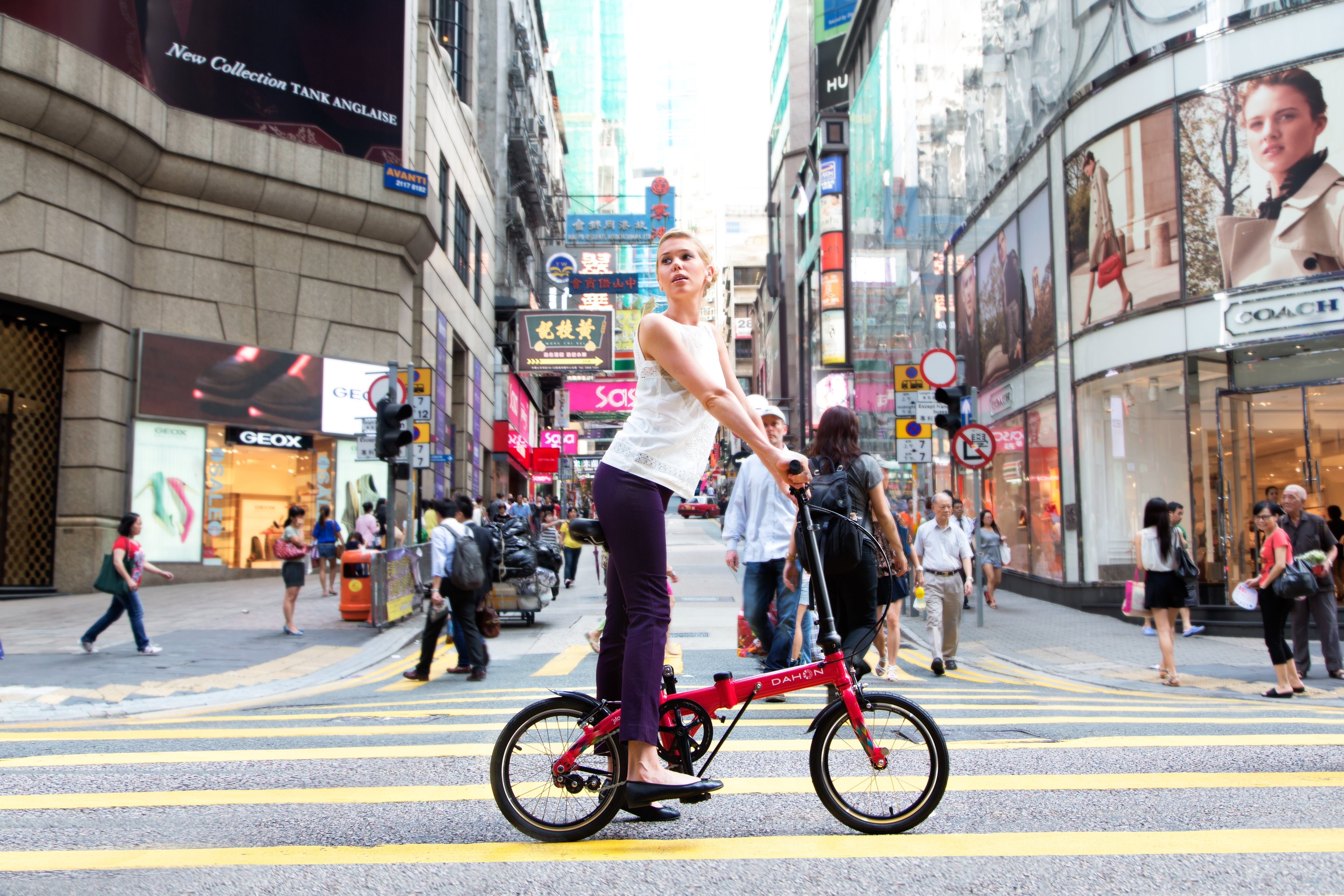 A woman rides a lightweight red folding bike in a busy city street, surrounded by pedestrians and storefronts.