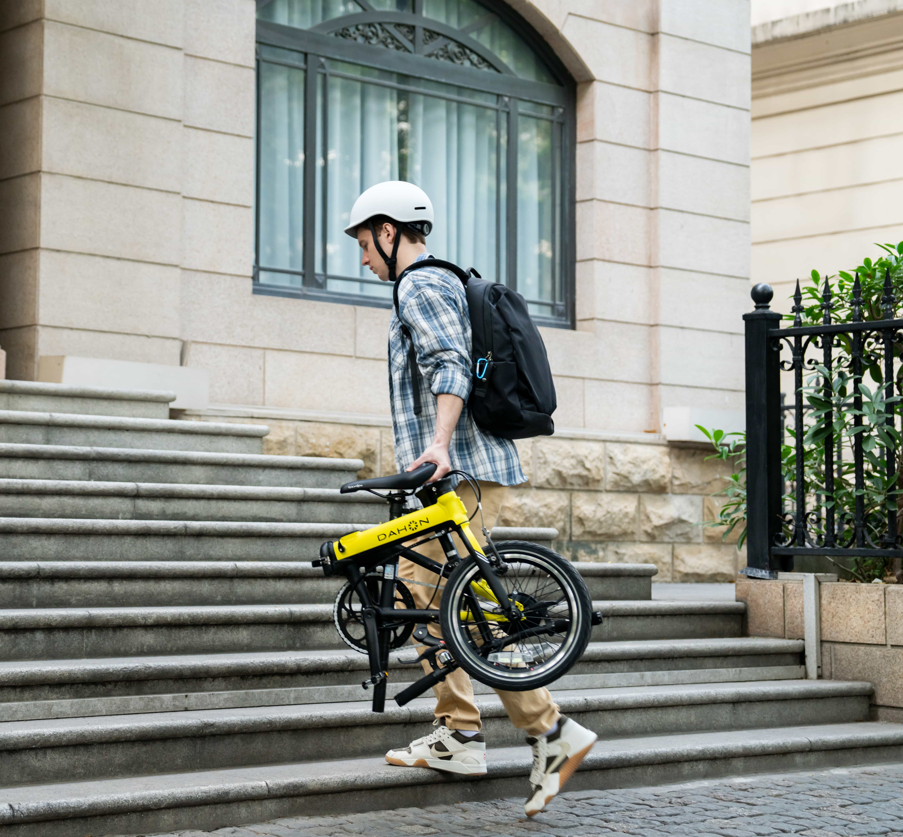 A man in casual attire carries a folded yellow DAHON bike up stone steps, showcasing its portability and urban versatility.