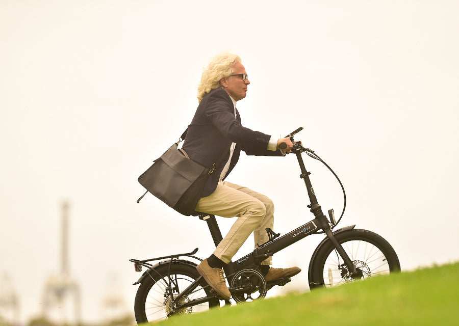Man with blond hair rides black DAHON Unio E20 foldable electric bike for urban commute, carrying messenger bag.