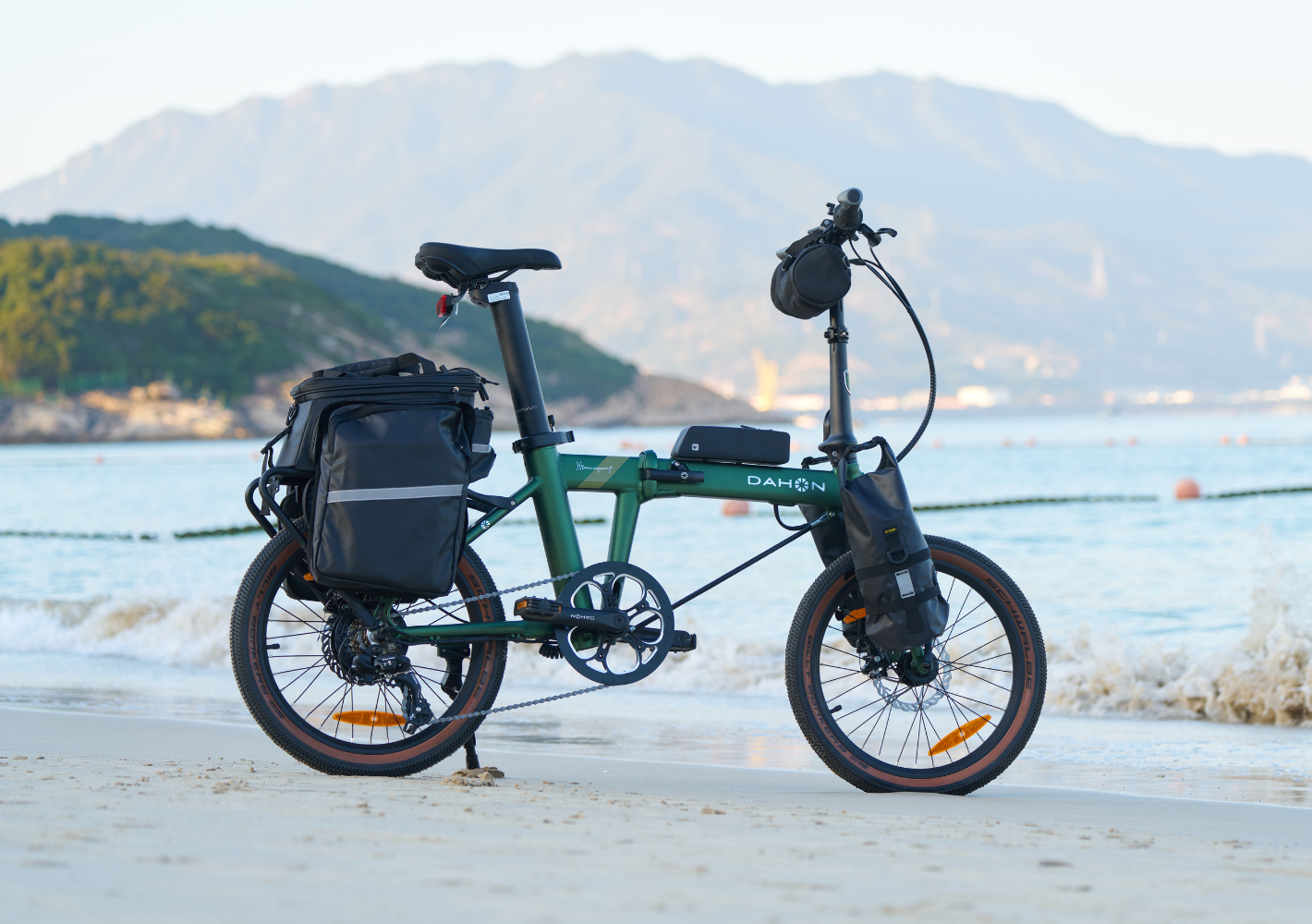 Deep green DAHON E-Hemingway foldable electric SUV bike on beach with saddlebags, front bag, and brown-rimmed tires.