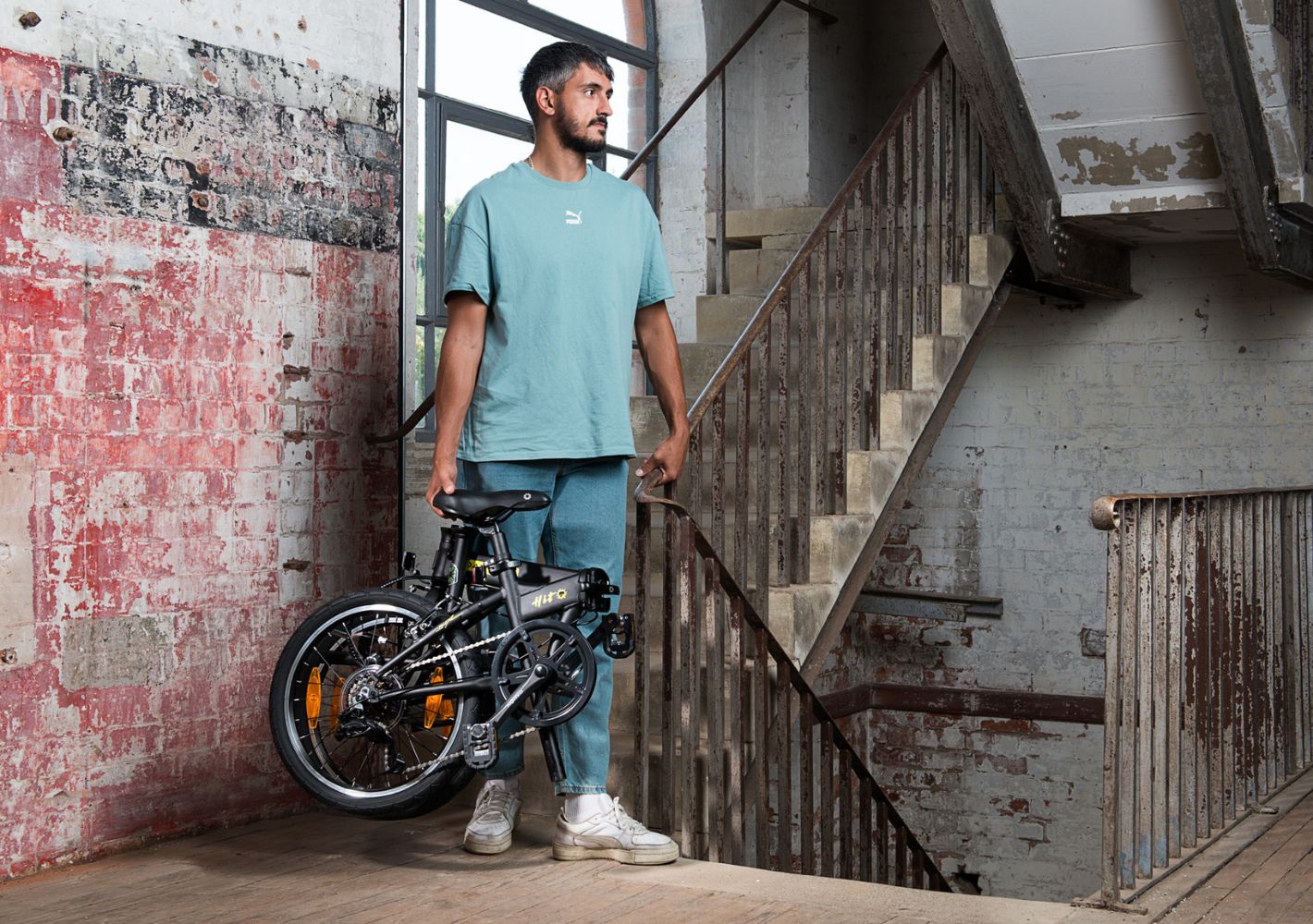 Man carries folded black Dahon HIT D6 folding bike with yellow accents in urban setting.
