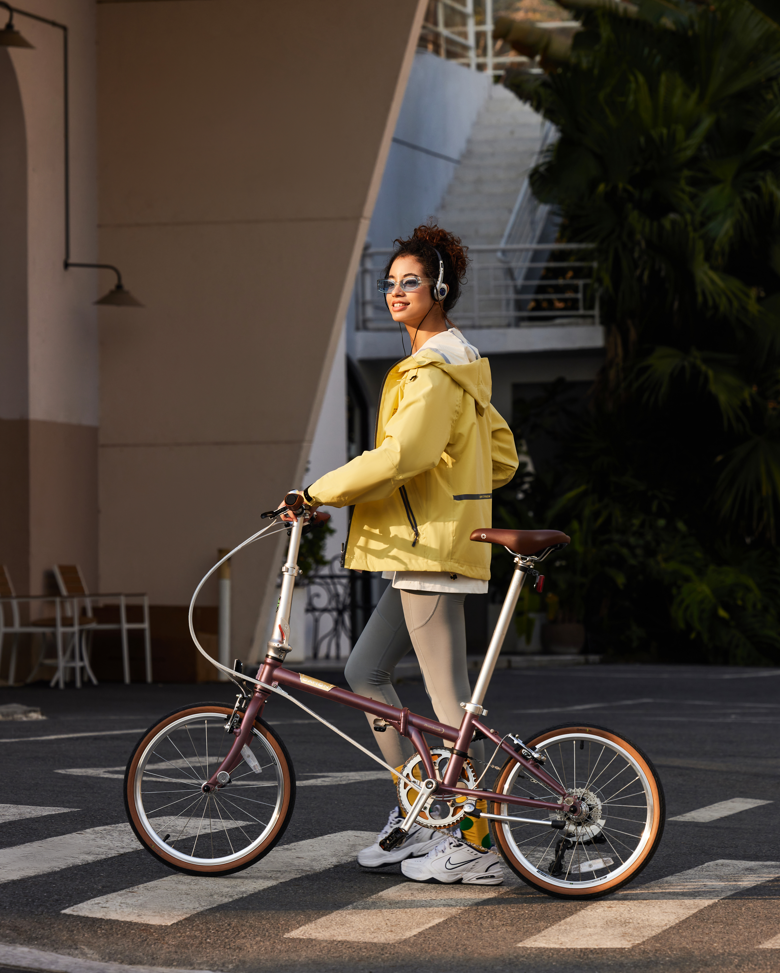 Woman in a yellow jacket with headphones walking a purple folding bike across a crosswalk, highlighting urban use.