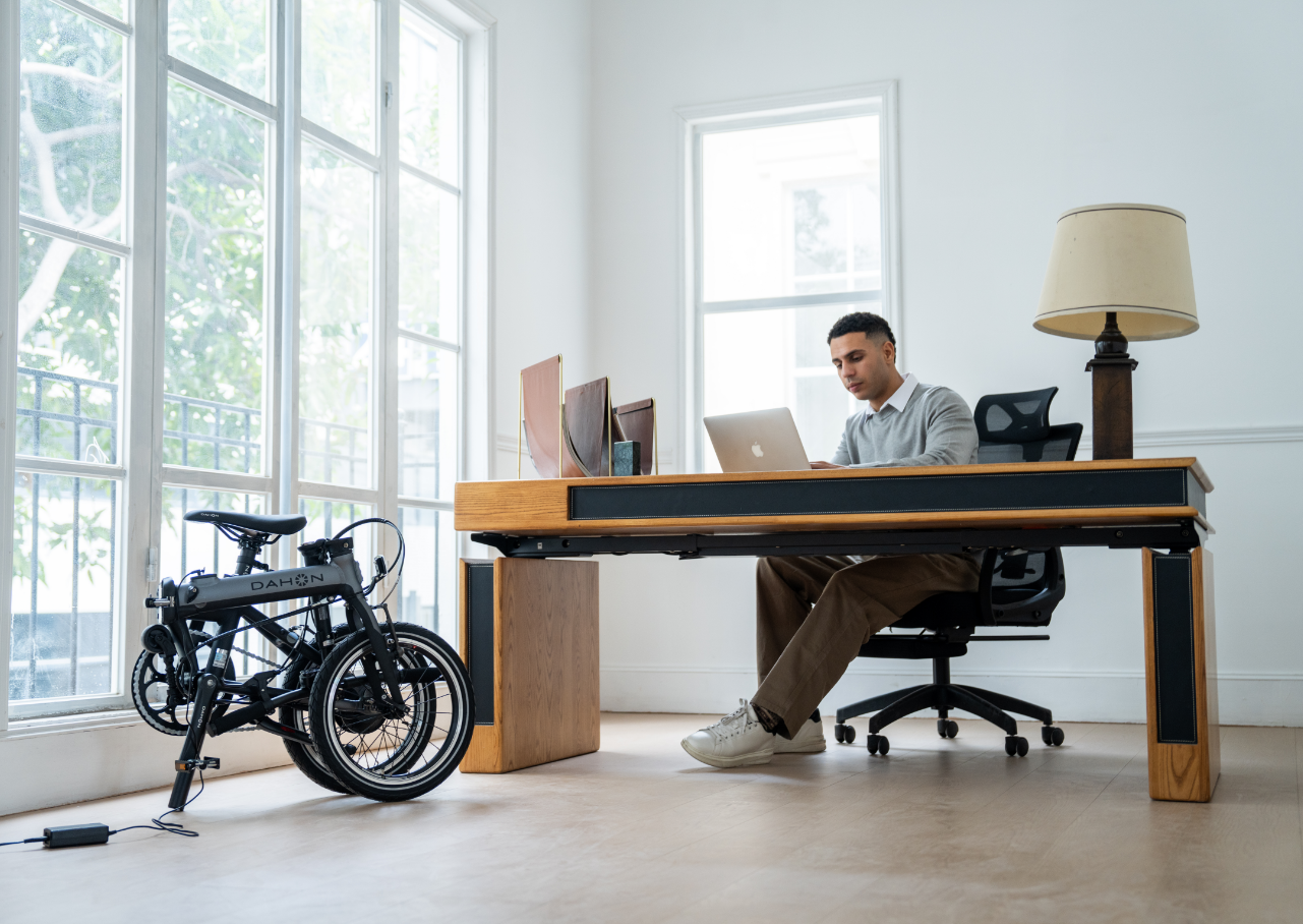 A person sitting at a modern desk in a bright, minimalist room, working on a laptop. A folding bicycle is parked next to the desk near large windows that let in natural light.