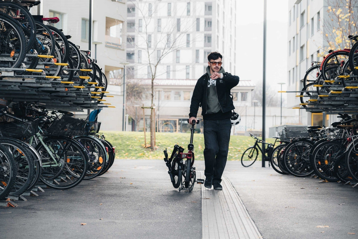 Man wheeling a folded bike through outdoor bicycle parking while checking his watch